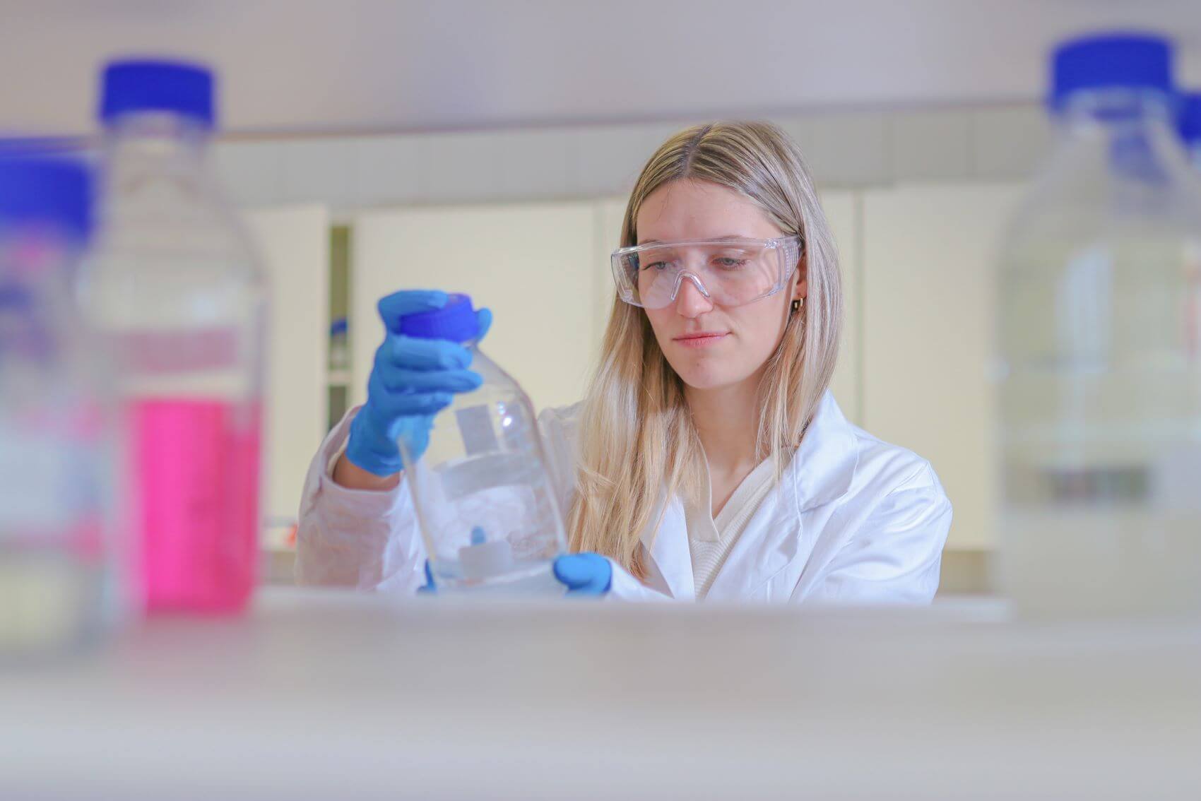 Foto von einer blonden Labormitarbeiterin. Sie trägt eine durchsichtige Schutzbrille und blaue Schutzhandschuhe. In der Hand hält sie eine Laborflasche