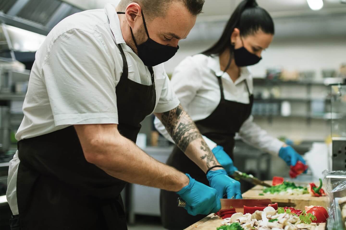 Zwei Köche mit Schutzmasken und Handschuhen schneiden frisches Gemüse in einer professionellen Restaurantküche – Hygienevorschriften in der Gastronomie.