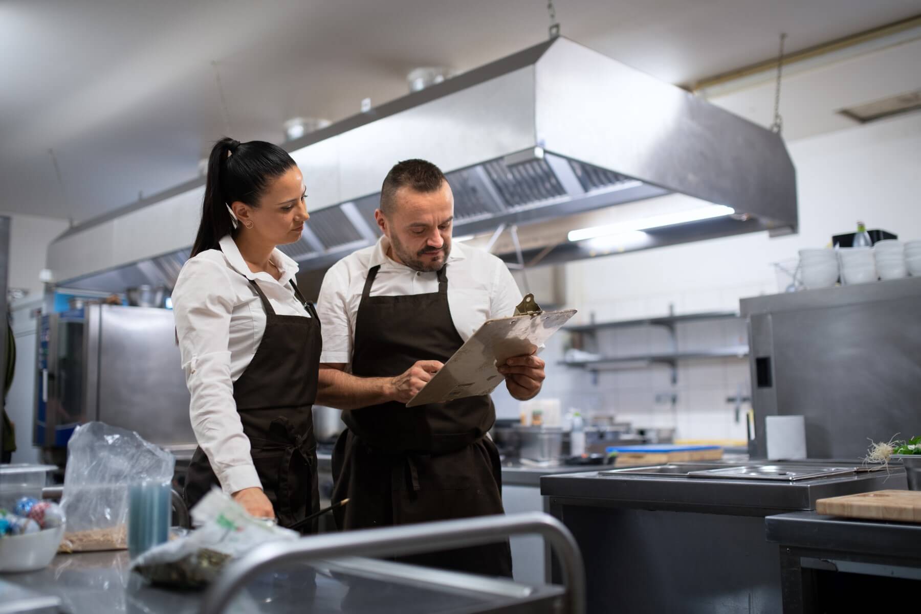Küchenpersonal überprüft einen Hygieneplan auf einem Klemmbrett in einer professionellen Gastronomieküche – Lebensmittelsicherheit und Qualitätskontrolle.
