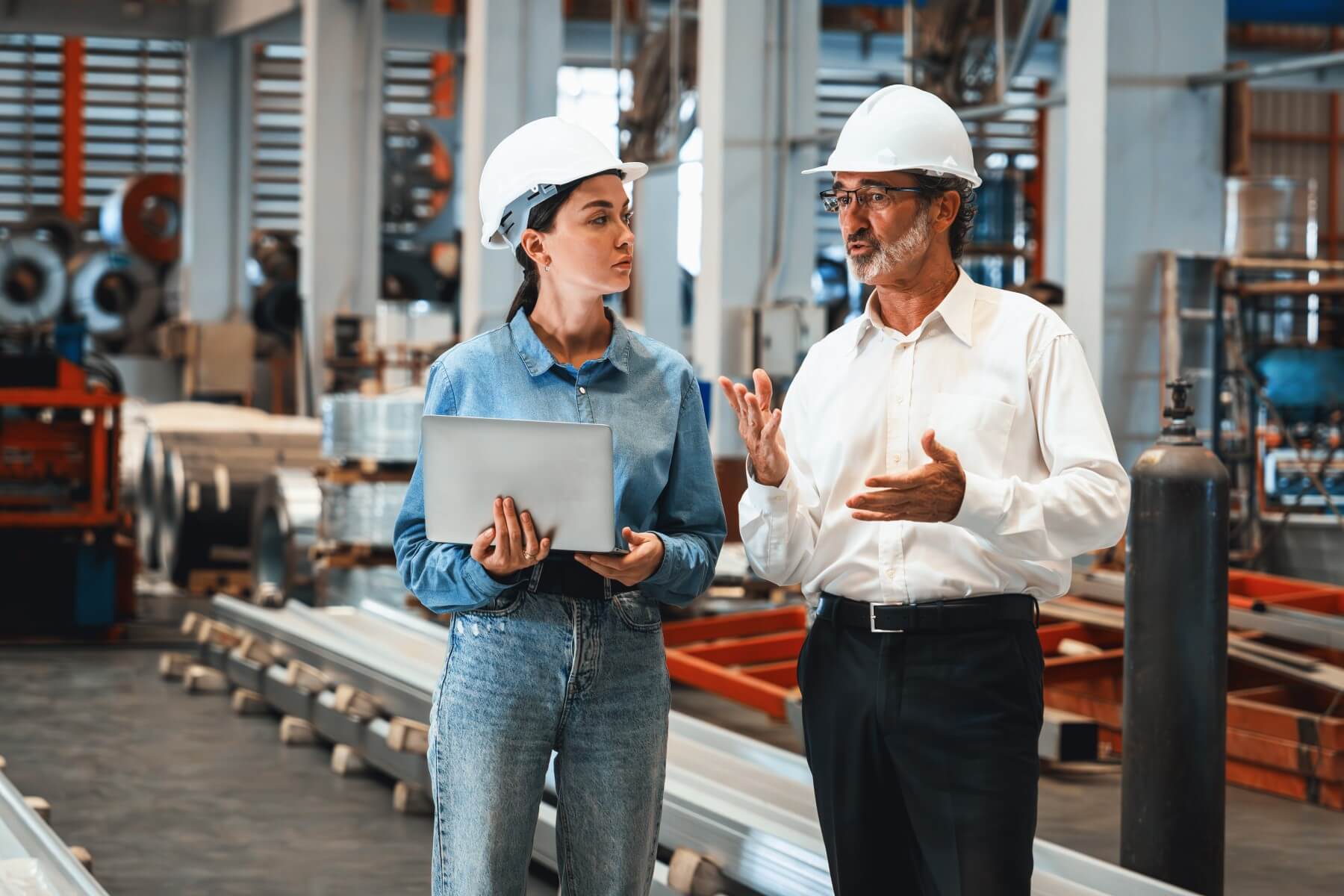 Produktionsleiter erklärt einer Ingenieurin mit Laptop Arbeitsprozesse in einer Industrieanlage mit Maschinen und Metallprofilen