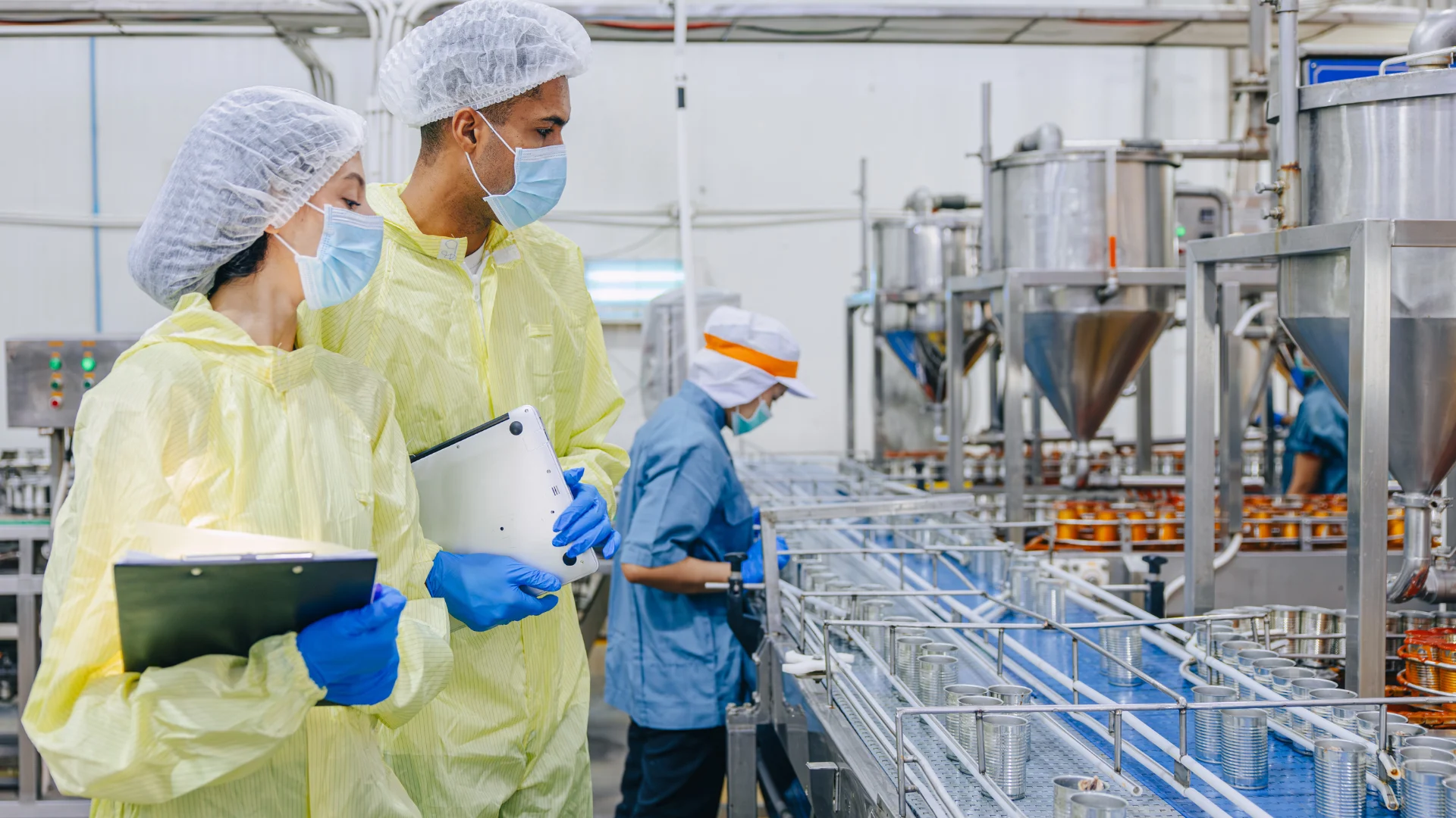 Zwei Personen in gelben Anzügen, Haarnetzen und blauen Handschuhen in einer Produktion